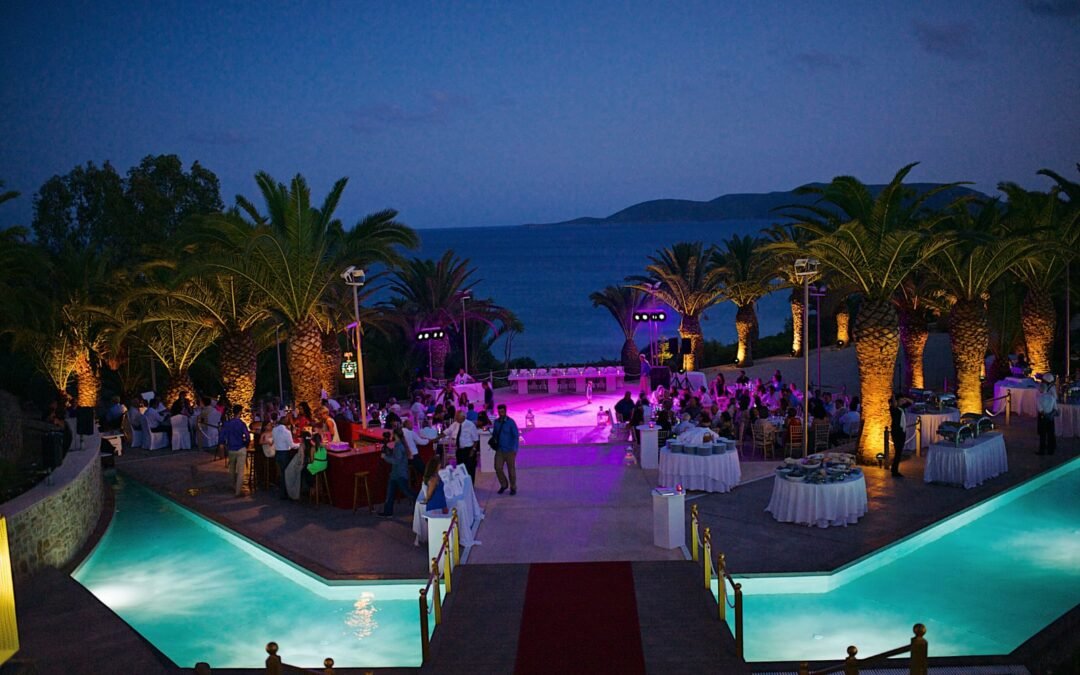 Evening wedding celebration at Stadion, Finikounda, Greece, on 31 July 2014 — guests dining and dancing under palm trees with glowing lights reflecting off two turquoise pools, overlooking the sea at sunset.