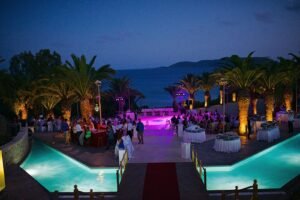 Evening wedding celebration at Stadion, Finikounda, Greece, on 31 July 2014 — guests dining and dancing under palm trees with glowing lights reflecting off two turquoise pools, overlooking the sea at sunset.