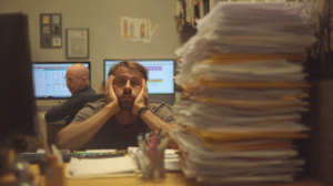 A small business owner looks overwhelmed by piles of process binders and paperwork while employees sit disengaged at their desks. The image captures how over-systemising kills initiative and traps leaders in daily chaos.