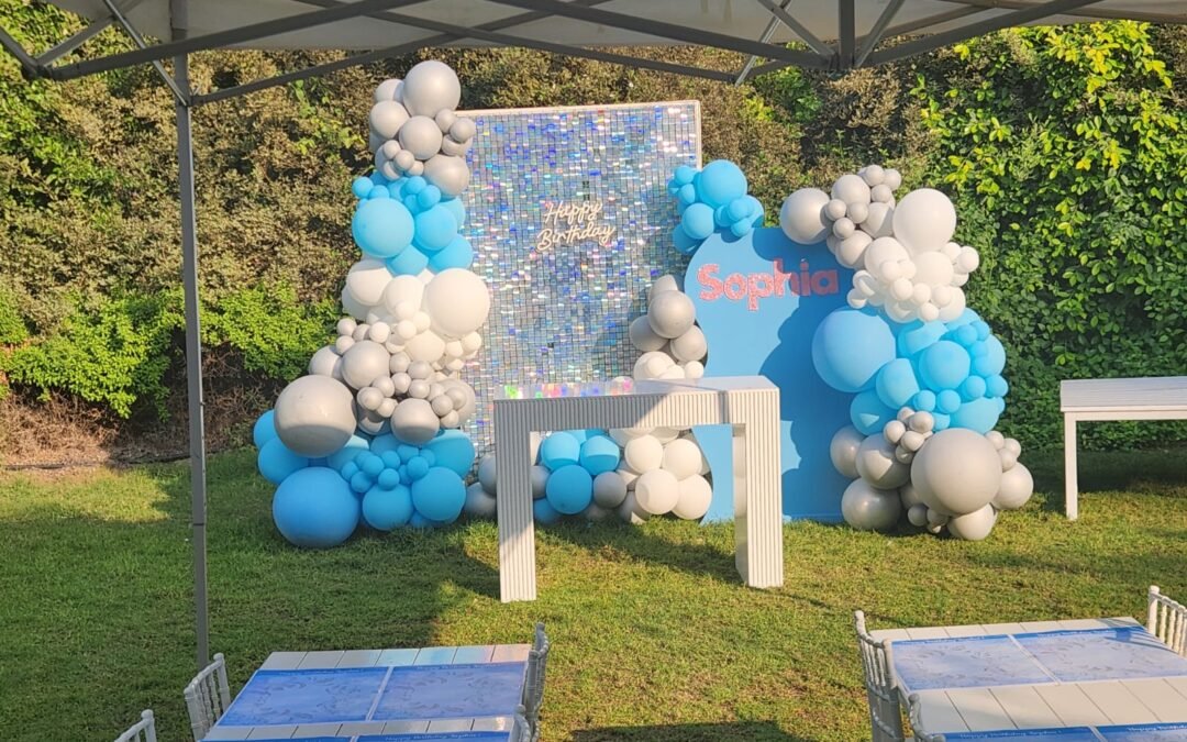 Blue white and silver balloons set up next to a sign saying happy 6th Birthday Sophia in front of green bushes on a beautiful sunny day