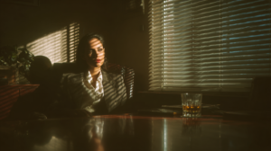 A businesswoman sitting alone in a dimly lit office, blinds casting sharp shadows across her face, a glass of whiskey on the desk, conveying tension and hard decisions