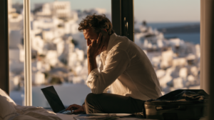 A weary businessman in his forties sitting on the edge of a hotel bed in a sunlit Greek island room, hunched over an open laptop with a phone pressed to his ear, an unopened suitcase beside him and whitewashed Santorini buildings and blue sea through the window behind, warm late afternoon light